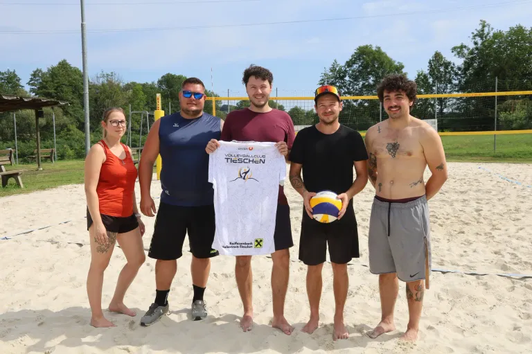 Fünf Personen stehen auf einem sandigen Strand, zwei Männer halten ein weißes Shirt mit einem Volleyball-Logo, ein Mann hält einen Volleyball und zwei Frauen stehen links.