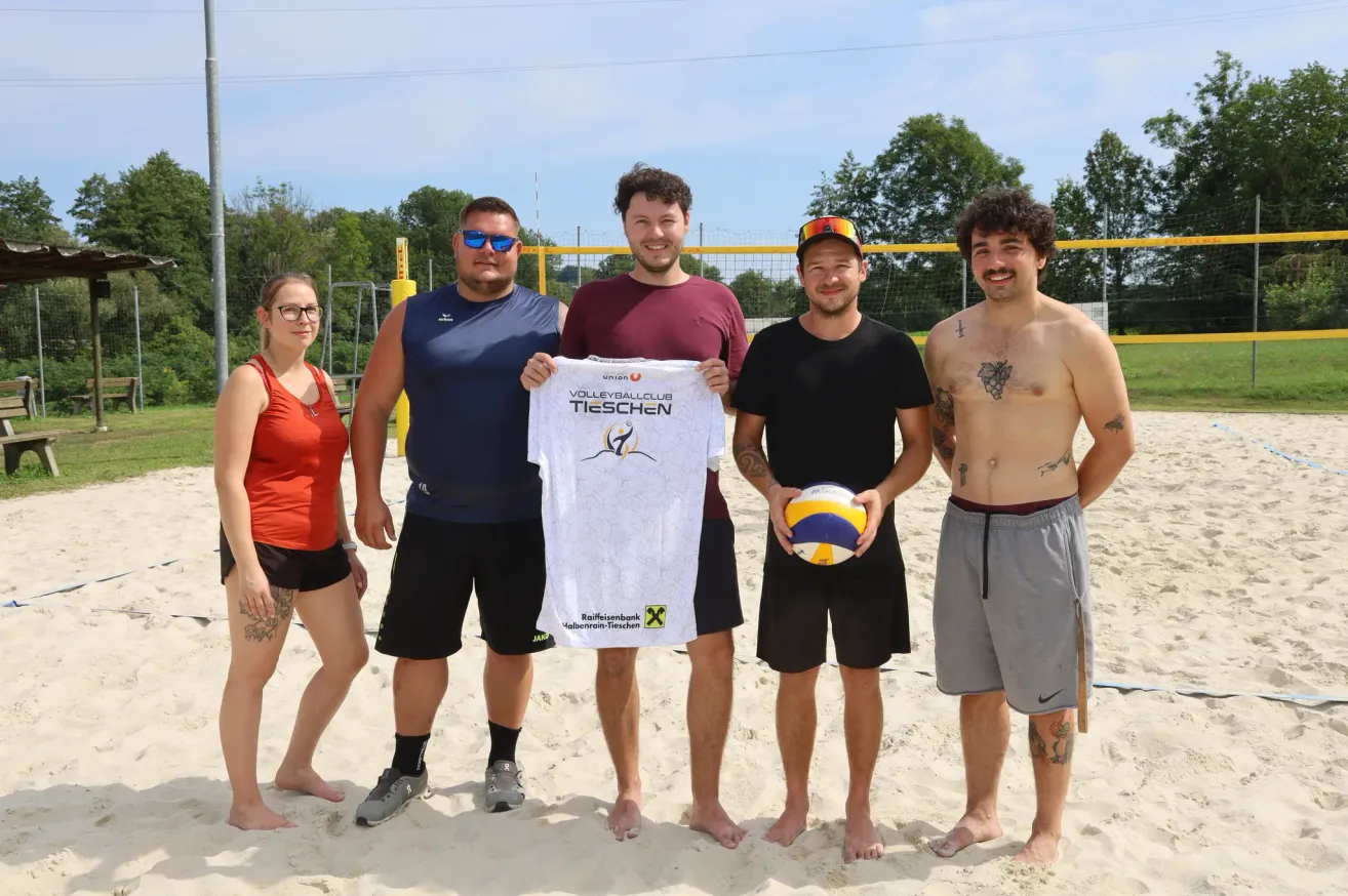 Fünf Personen stehen auf einem sandigen Strand, zwei Männer halten ein weißes Shirt mit einem Volleyball-Logo, ein Mann hält einen Volleyball und zwei Frauen stehen links.