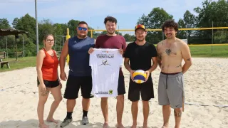 Fünf Personen stehen auf einem sandigen Strand, zwei Männer halten ein weißes Shirt mit einem Volleyball-Logo, ein Mann hält einen Volleyball und zwei Frauen stehen links.