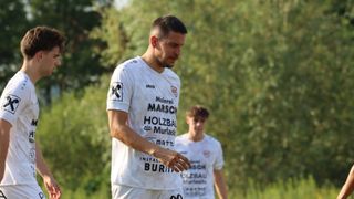 A soccer player in a white jersey with the word 'Malere' on it is looking down while walking on a field. Another player in a similar jersey is in the background.
