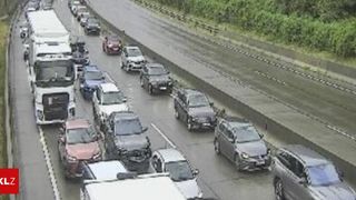 A highway with multiple lanes of traffic, including cars and trucks, moving slowly in the left lane. The road is wet, and there are green vegetation and metal barriers on the side.