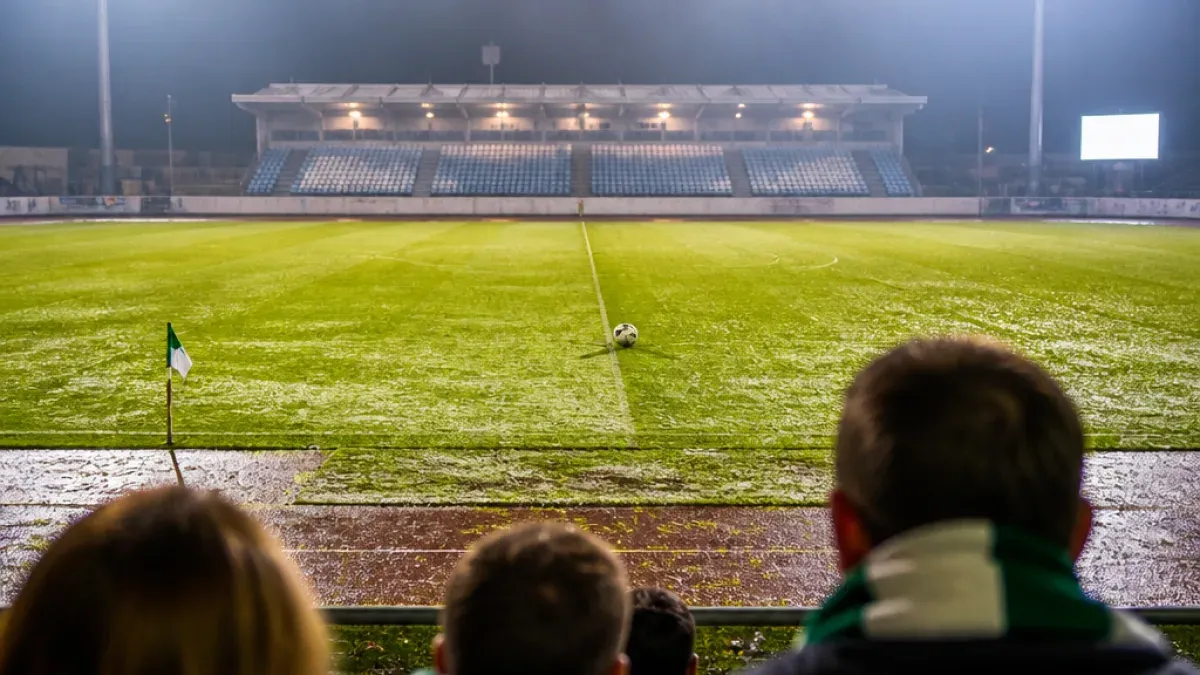 A nighttime soccer game in progress, with a soccer ball on the field and spectators watching from the stands.