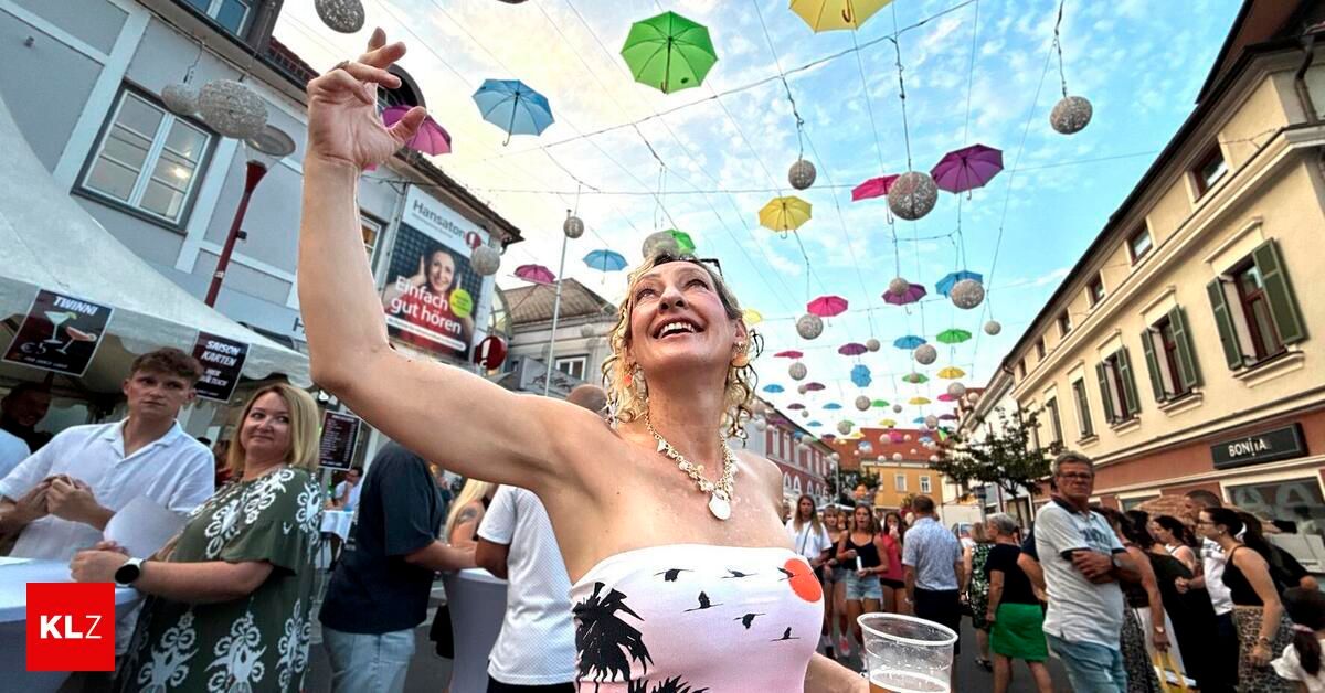 A woman in a pink top is smiling and raising her hand in a crowded street with colorful umbrellas hanging from the sky.