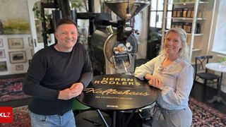 A man and a woman stand beside a round table with the words 'Herrn Rodlers Kaffeehaus' in front of a coffee roaster.