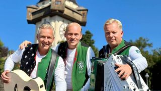 Three men dressed in traditional green attire are standing in front of a clock tower, smiling and posing for a photo. The man on the left holds a guitar, the middle one plays an accordion, and the right one has a microphone.