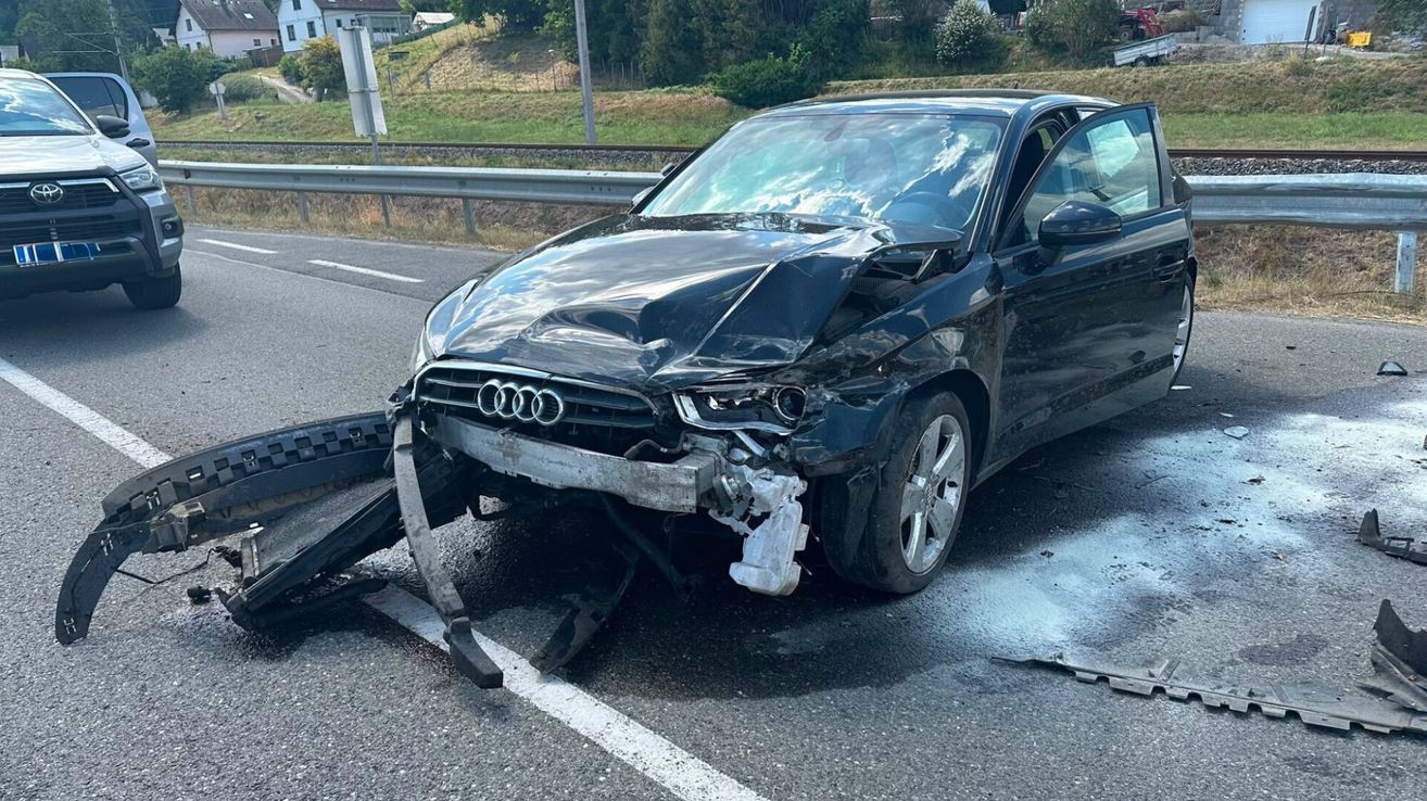 A black Audi car with severe front-end damage is parked on the side of a road. The bumper is detached, and the hood is crumpled.