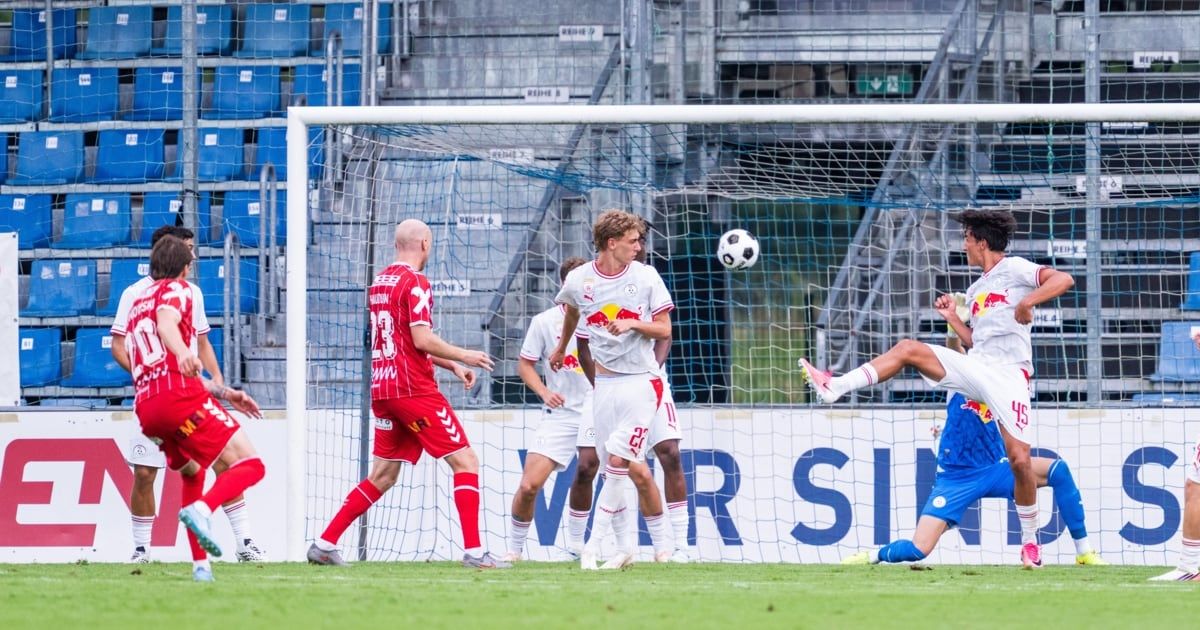 Soccer players from two teams compete for the ball in front of the goal. The player in white is attempting to block the shot.