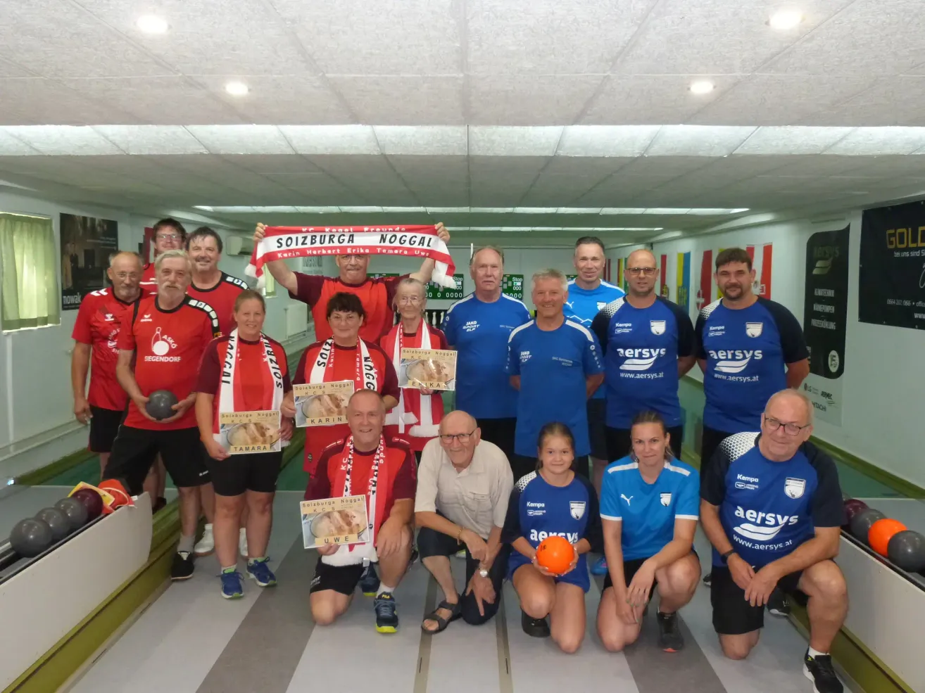A group of people dressed in red and blue jerseys, some holding trophies, posing for a photo in a sports hall.