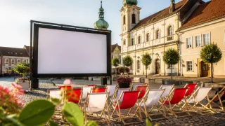 A large outdoor movie screen is set up in front of a historic building with a green dome. Rows of red and white folding chairs are arranged in front of the screen.