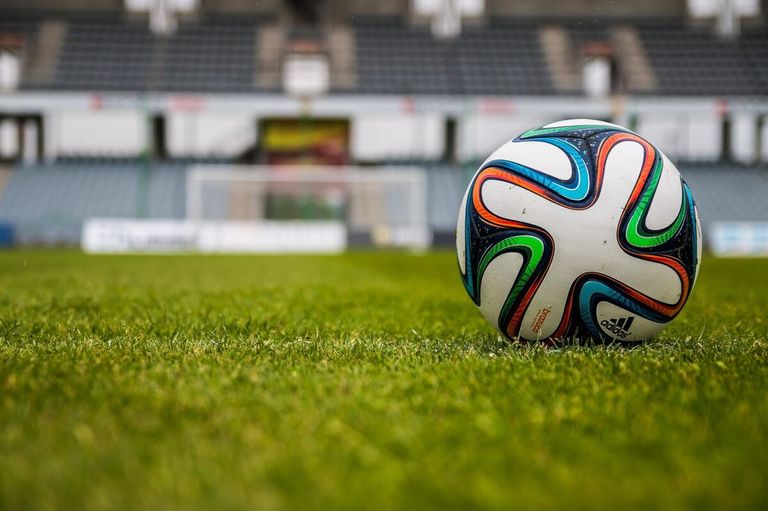 A soccer ball with Adidas branding sits on a lush green field, with a blurry background of an empty stadium in the distance.