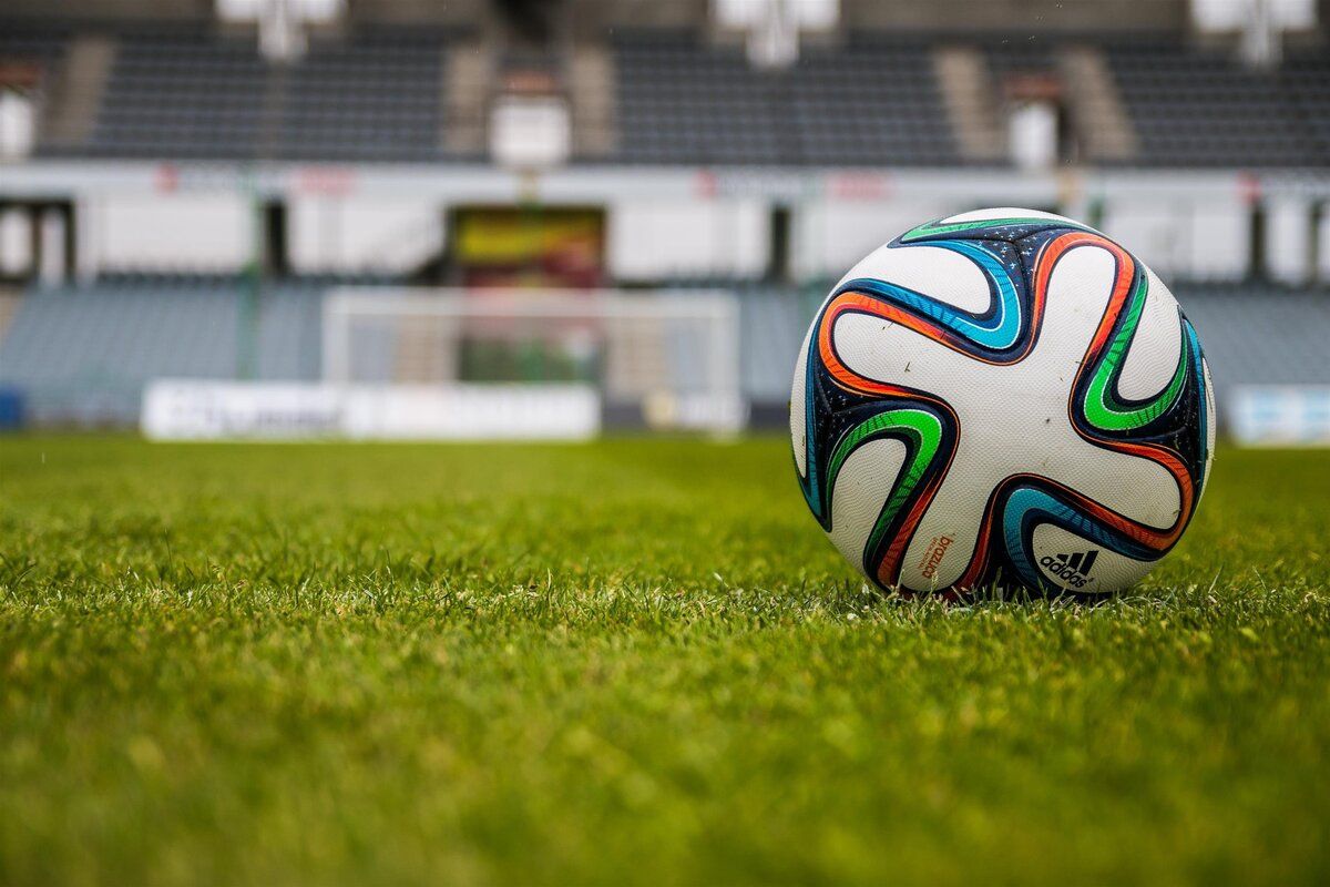 A soccer ball with Adidas branding sits on a lush green field, with a blurry background of an empty stadium in the distance.