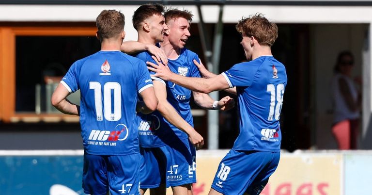 A group of soccer players in blue uniforms celebrate a goal on a field. The player in the middle has his mouth open and his arms around the shoulders of two other players.