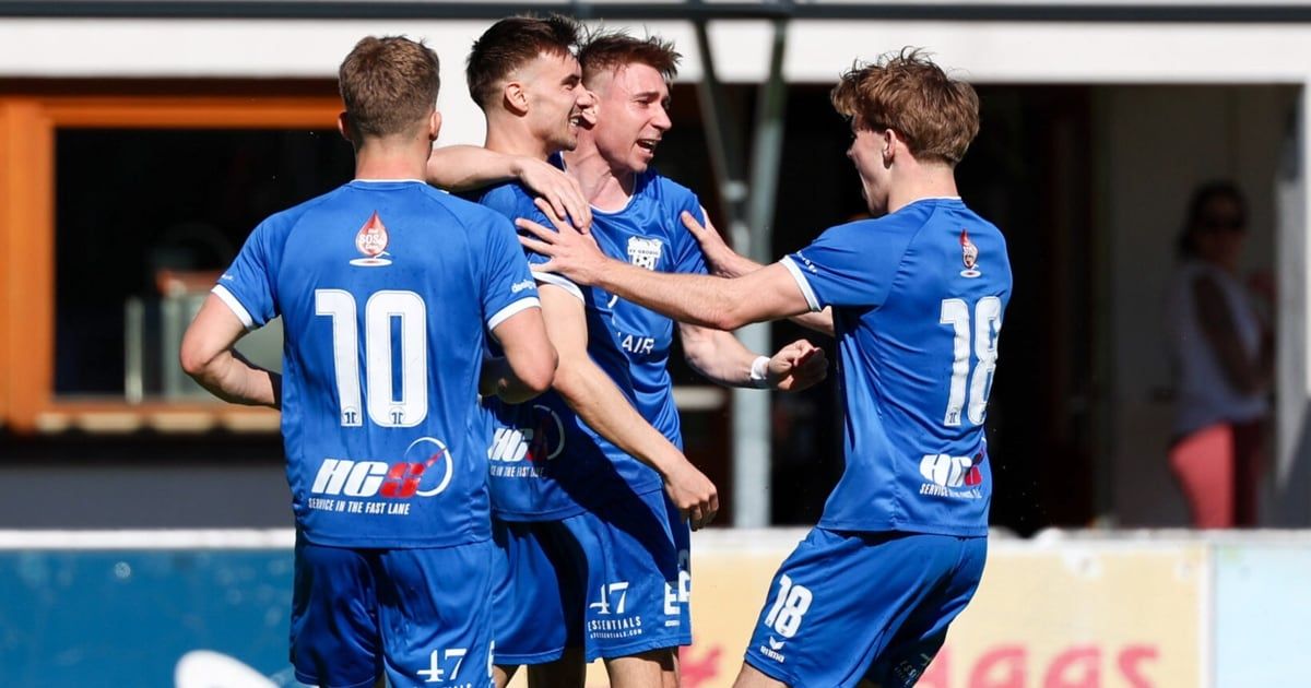 Eine Gruppe von Fußballspielern in blauen Trikots feiert ein Tor auf einem Feld. Der Spieler in der Mitte hat den Mund offen und seine Arme um die Schultern zweier anderer Spieler gelegt.