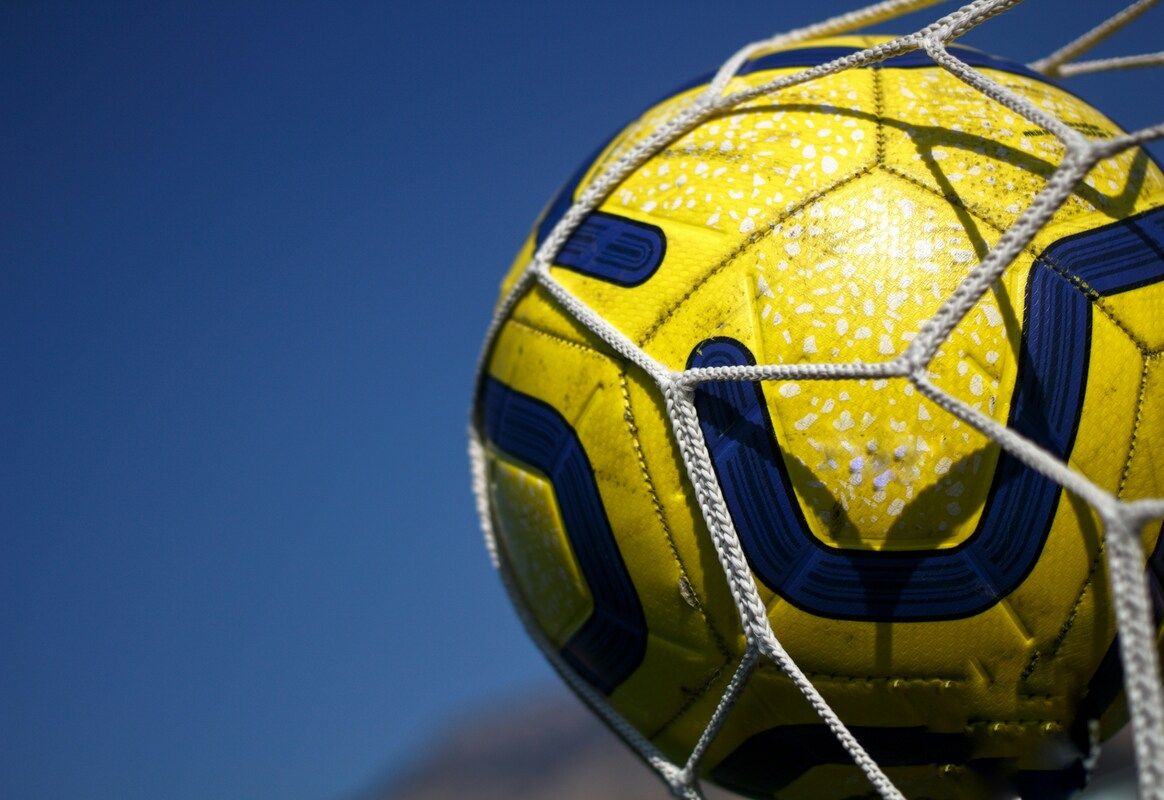 Eine Nahaufnahme eines gelben Fußballs mit blauen Akzenten und weißen Sprenkeln, teilweise in einem weißen Netz gefangen, vor einem blauen Himmel.