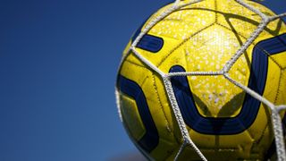 A close-up of a yellow soccer ball with blue accents, partially caught in a white net against a blue sky.