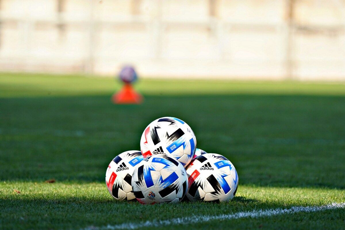 Five Adidas soccer balls are stacked on a grassy field with a blurry background showing a white building and a person in the distance.