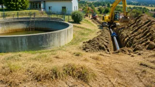 Eine Baustelle mit einem gelben Bagger, der eine Grube gräbt, und einem großen Betonwassertank in der Nähe. Im Hintergrund sind ein weißes Haus und ein weiterer Bagger zu sehen.