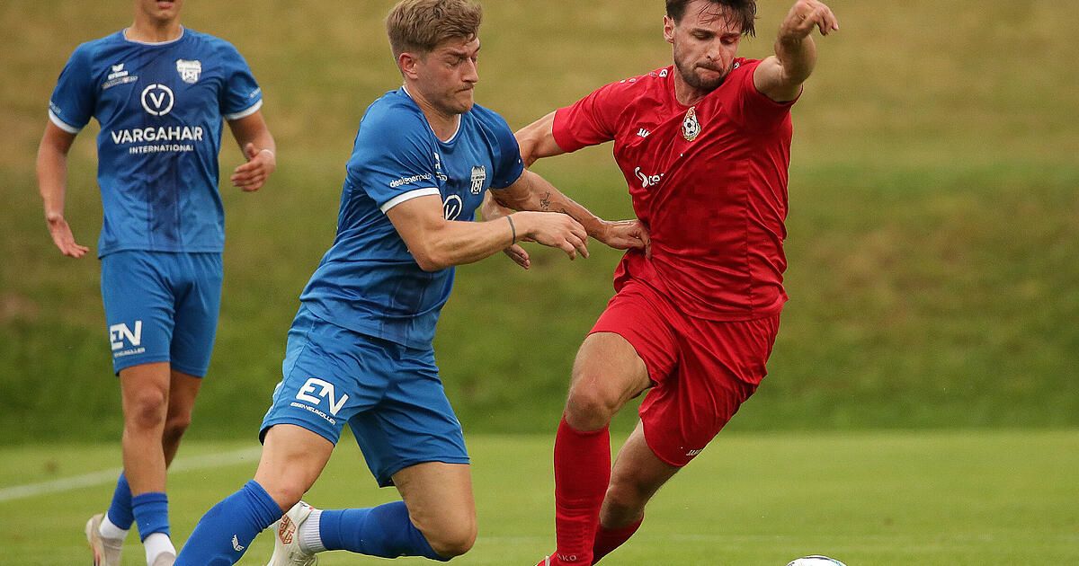 Zwei Fußballspieler, einer in Blau und einer in Rot, kämpfen auf einem Rasenfeld um den Ball.