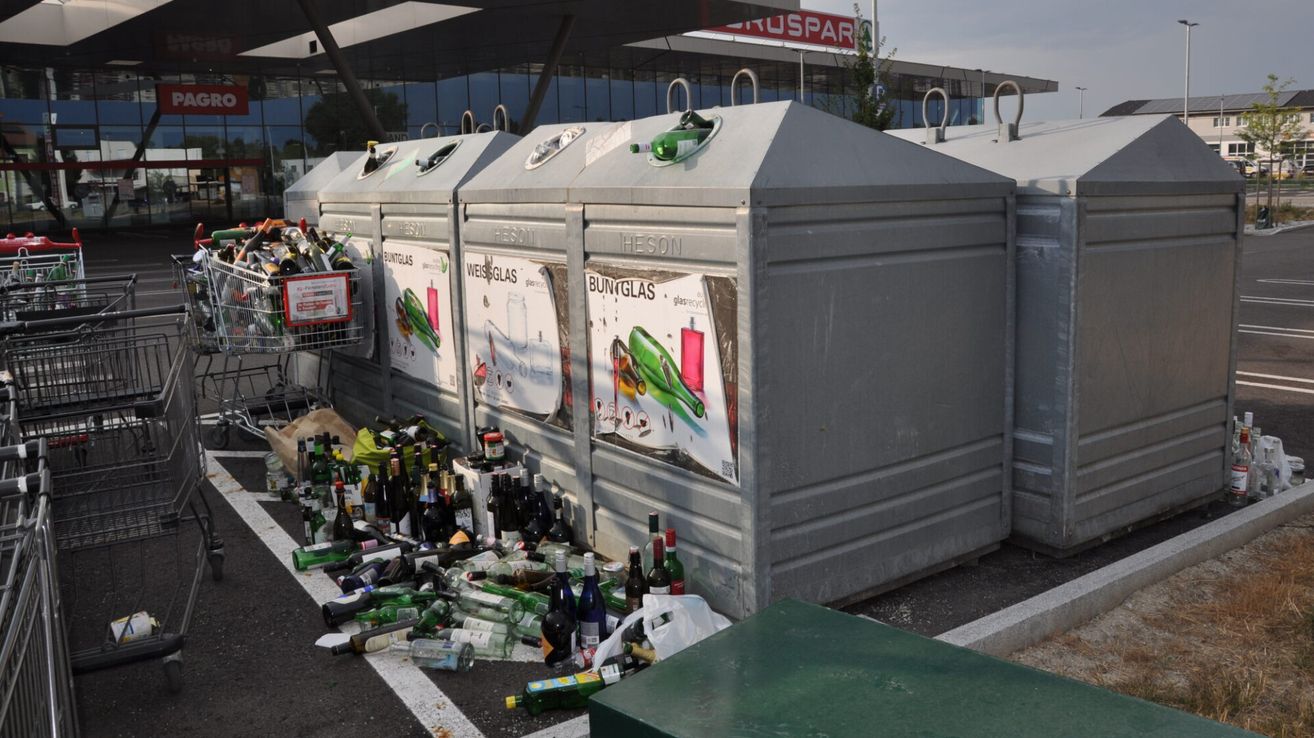 Eine Reihe von grauen Recyclingbehältern mit Etiketten für verschiedene Abfallarten, einschließlich Glas, ist von einem Haufen leerer Flaschen und anderem Müll umgeben.