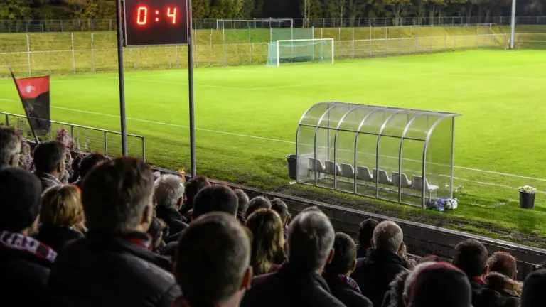 Ein Fußballfeld mit einer Anzeigetafel, die 0:4 anzeigt, eine Gruppe von Menschen, die von den Tribünen zuschauen, und eine leere Glasunterkunft mit Stühlen.