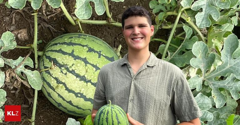 Ein junger Mann steht in einem Garten und hält eine Wassermelone in den Händen. Er lächelt und hinter ihm steht eine große Wassermelone.