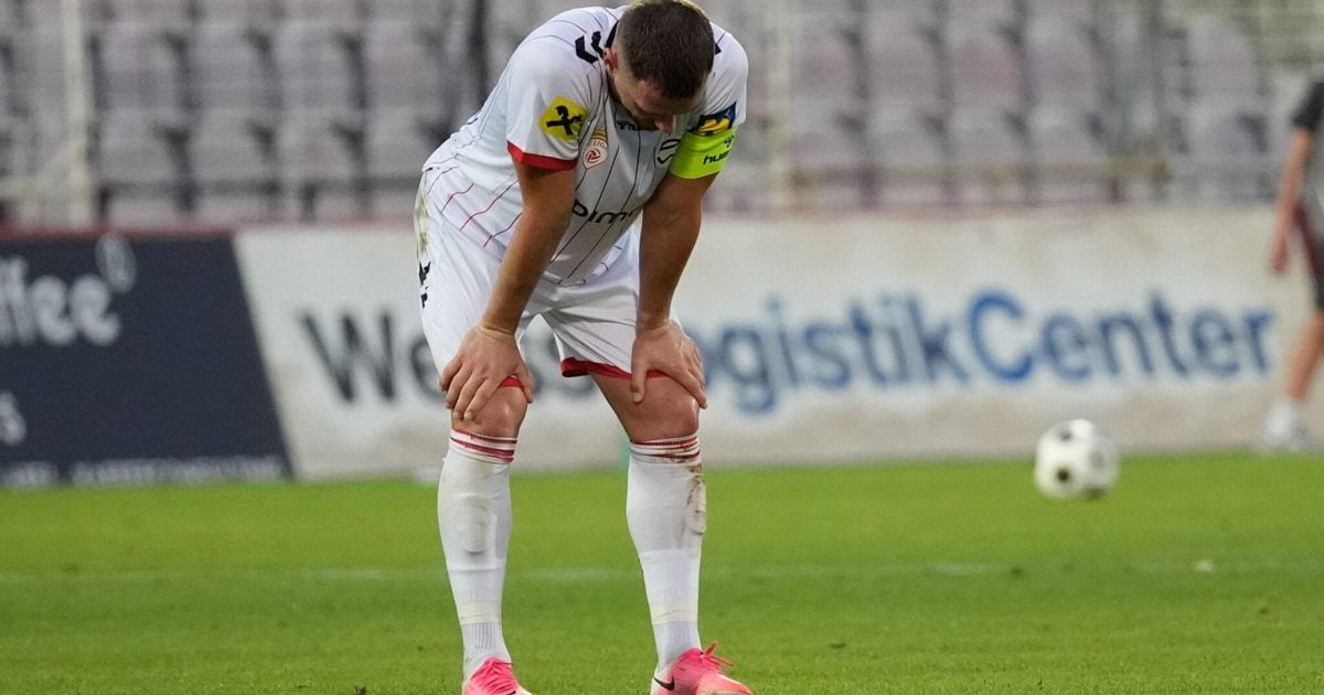 Ein Fußballspieler in einem weißen Trikot mit roten Akzenten ist vor Schmerz über sein Knie gebeugt auf einem Fußballfeld. Im Hintergrund ist ein weißes Banner mit blauem Text zu sehen.
