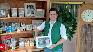 Ein Mann in einem grünen Weste hält ein Schwarz-Weiß-Foto vor einem Bücherregal mit verschiedenen Gegenständen.