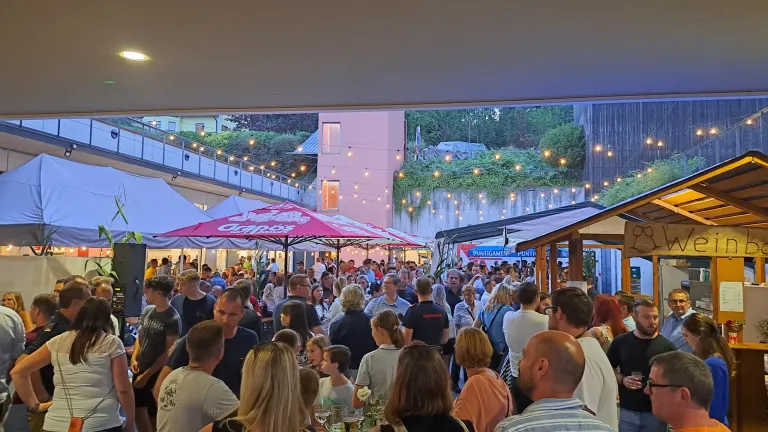 Eine überfüllte Outdoor-Veranstaltung mit Menschen unter Schirmen und Zelten, beleuchtet von Lichterketten und umgeben von Grün.