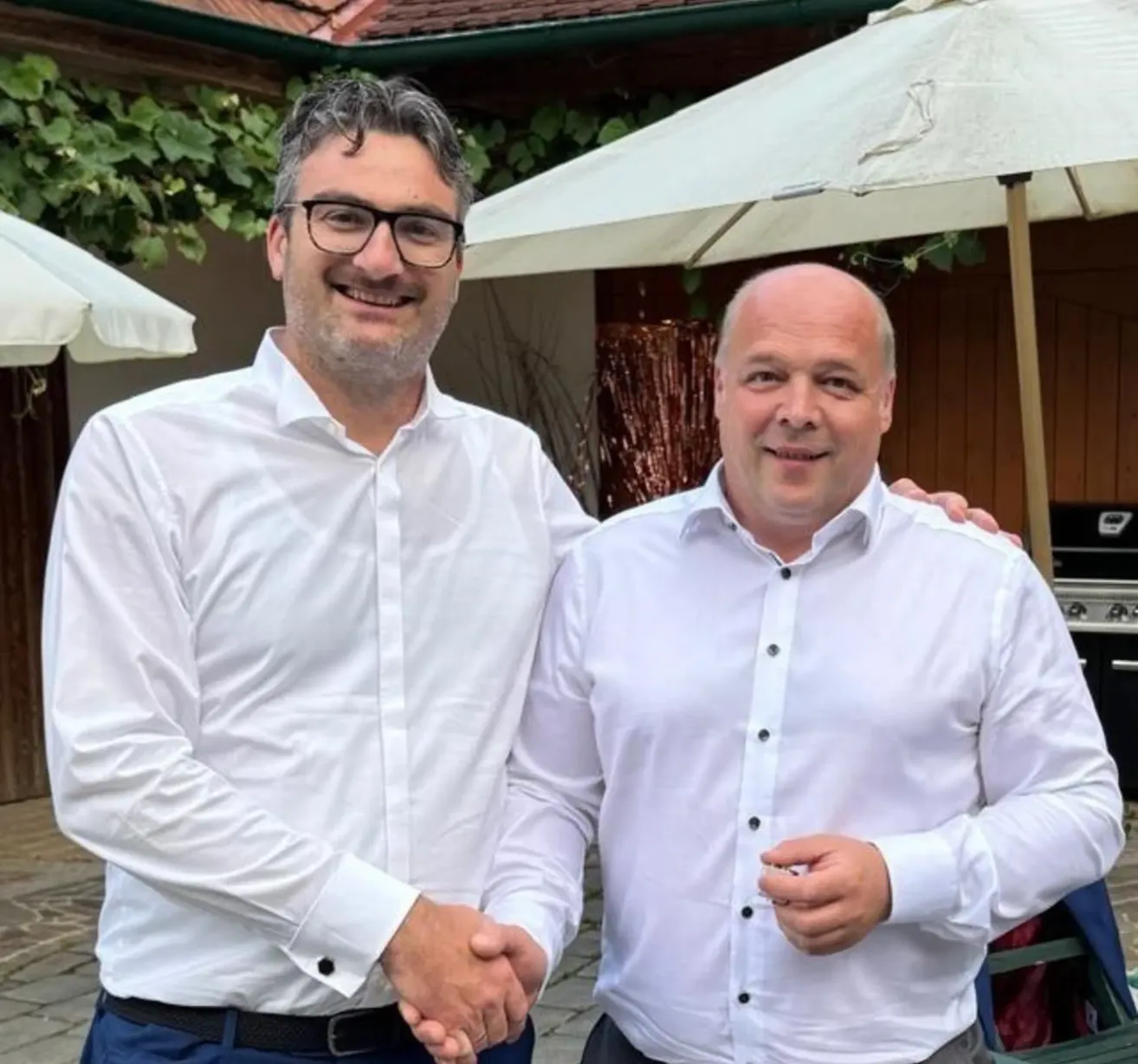 Zwei Männer in weißen Hemden und mit Brille lächeln und schütteln sich die Hände vor einem Schirm.
