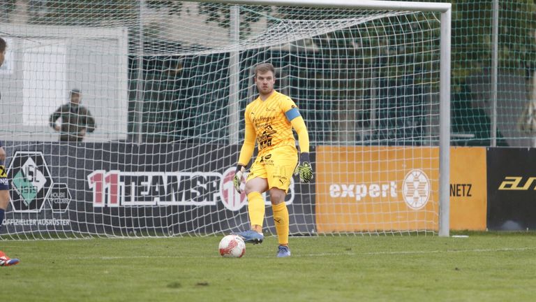 Ein Fußballspieler in einem gelben Trikot ist dabei, einen Ball auf einem Feld zu schießen, mit einem Tornetz im Hintergrund.