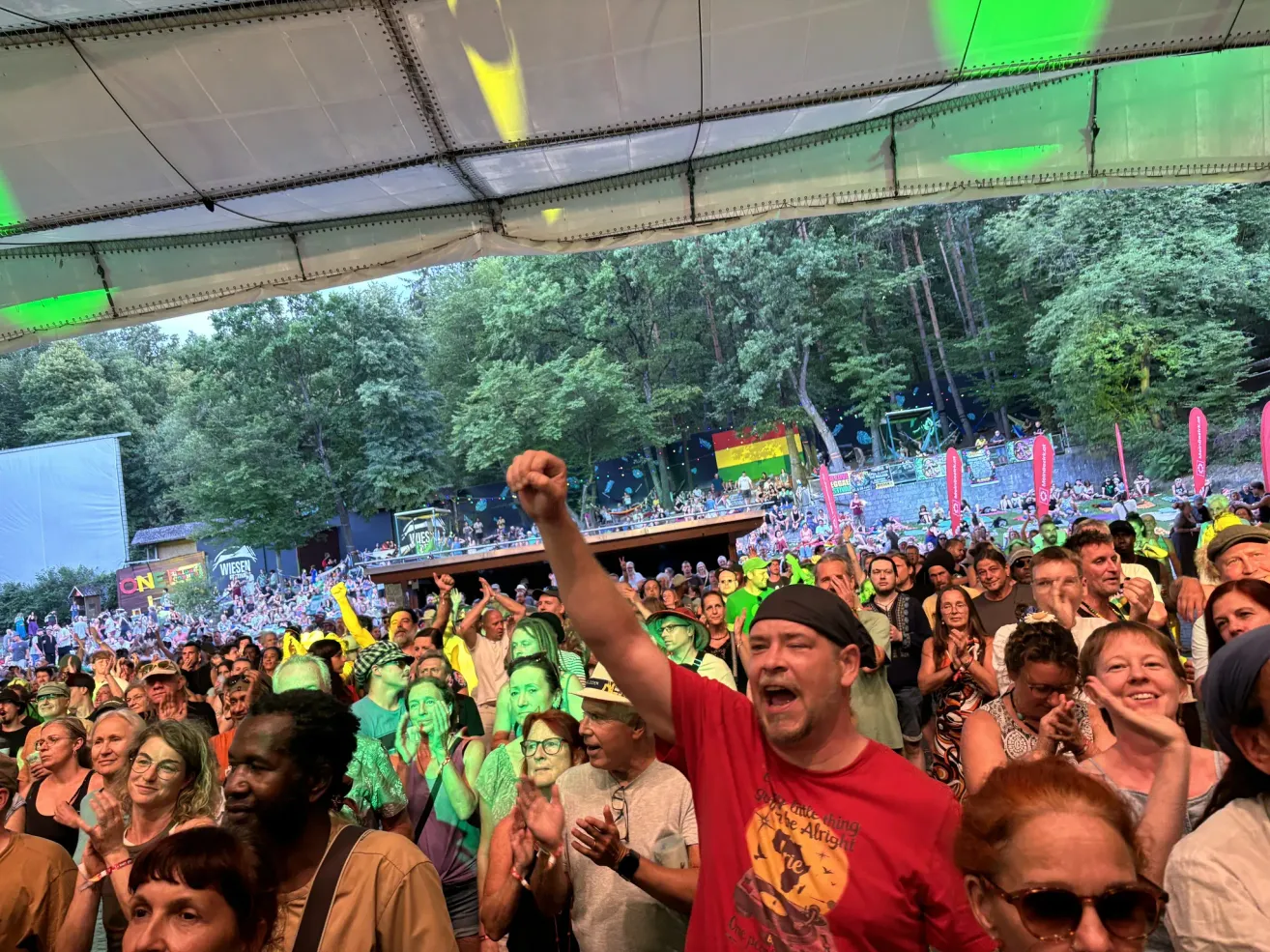 Eine große Menschenmenge bei einem Open-Air-Musikfestival, einige jubeln und andere tanzen, unter einem großen Zelt mit Bäumen im Hintergrund.