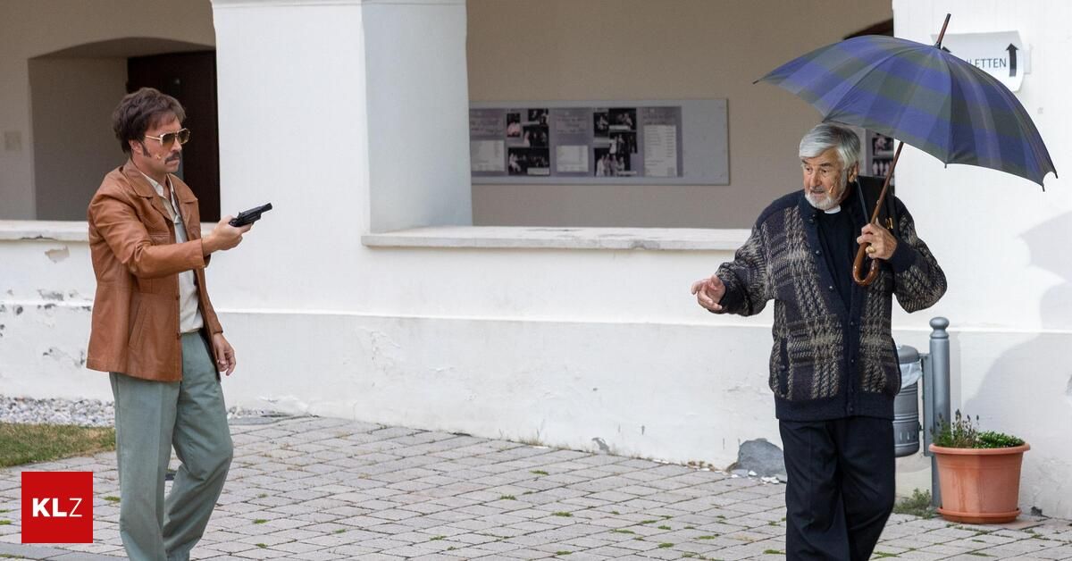Ein älterer Mann mit einer karierten Jacke und einem Regenschirm gestikuliert, während eine andere Person eine Waffe vor einem Gebäude hält.