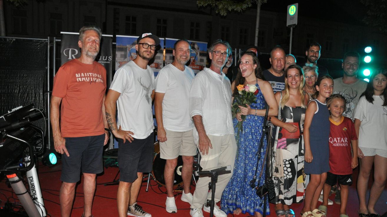 Eine Gruppe von Menschen steht auf einem roten Teppich und lächelt für ein Foto. Eine Frau hält einen Blumenstrauß. Hinter ihnen sind ein Schlagzeug und ein Mikrofon zu sehen.