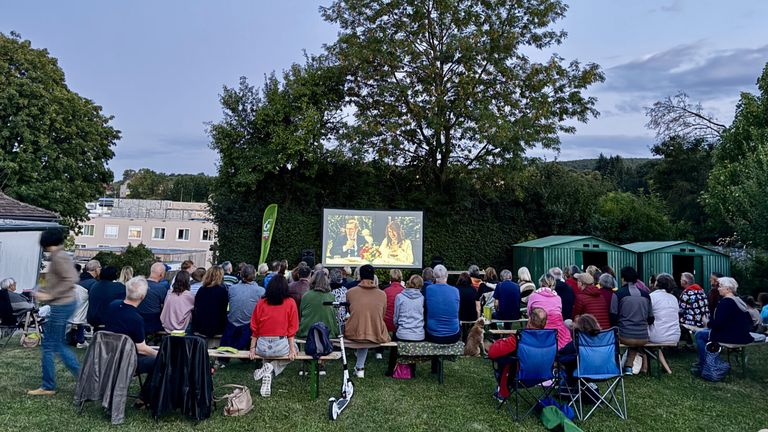 Eine Gruppe von Menschen versammelt sich im Freien, um einen Film auf einer Leinwand zu sehen. Die Leinwand zeigt einen Mann und eine Frau.