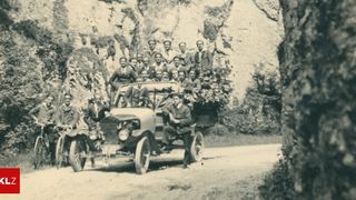 Ein Vintage-Foto zeigt eine Gruppe von Männern in formeller Kleidung, die in einem alten Auto fahren, wobei einige auf der Rückseite stehen und andere im Inneren sitzen, alle lächeln sie für die Kamera.