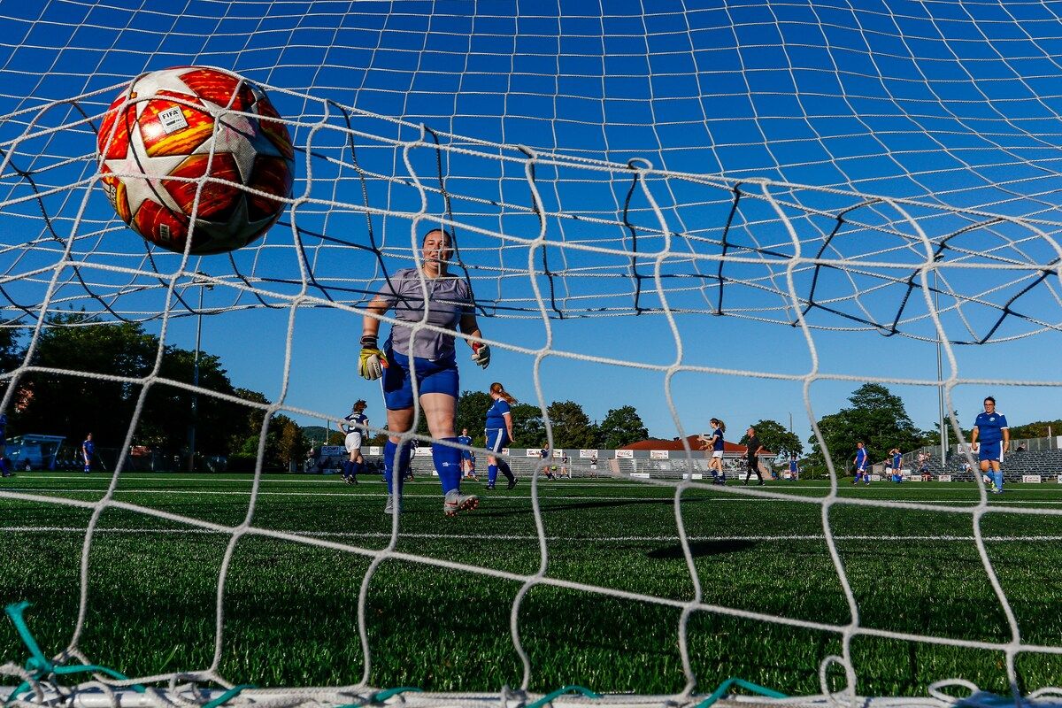 Ein Fußballspieler steht vor dem Tornetz, mit einem Fußball in der Luft, und anderen Spielern im Hintergrund.