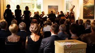 Eine Gruppe von Menschen sieht ein klassisches Musikkonzert mit Cello und Gemälde an der Wand.