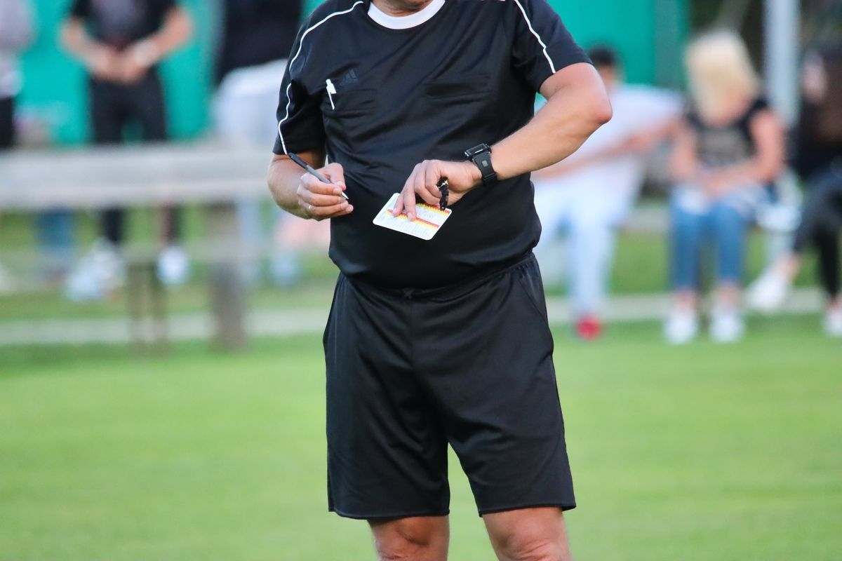 Ein Fußballschiedsrichter steht auf einem Feld, hält eine Karte und einen Stift in der Hand, mit einem verschwommenen Hintergrund von Spielern und Zuschauern.