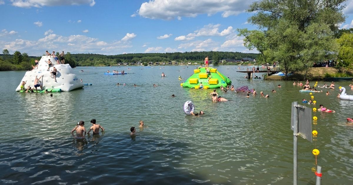 Eine große Gruppe von Menschen genießt einen sonnigen Tag an einem See mit aufblasbaren Wasserspielzeug und einer Wasserrutsche. Bäume und ein entferntes Ufer sind sichtbar.