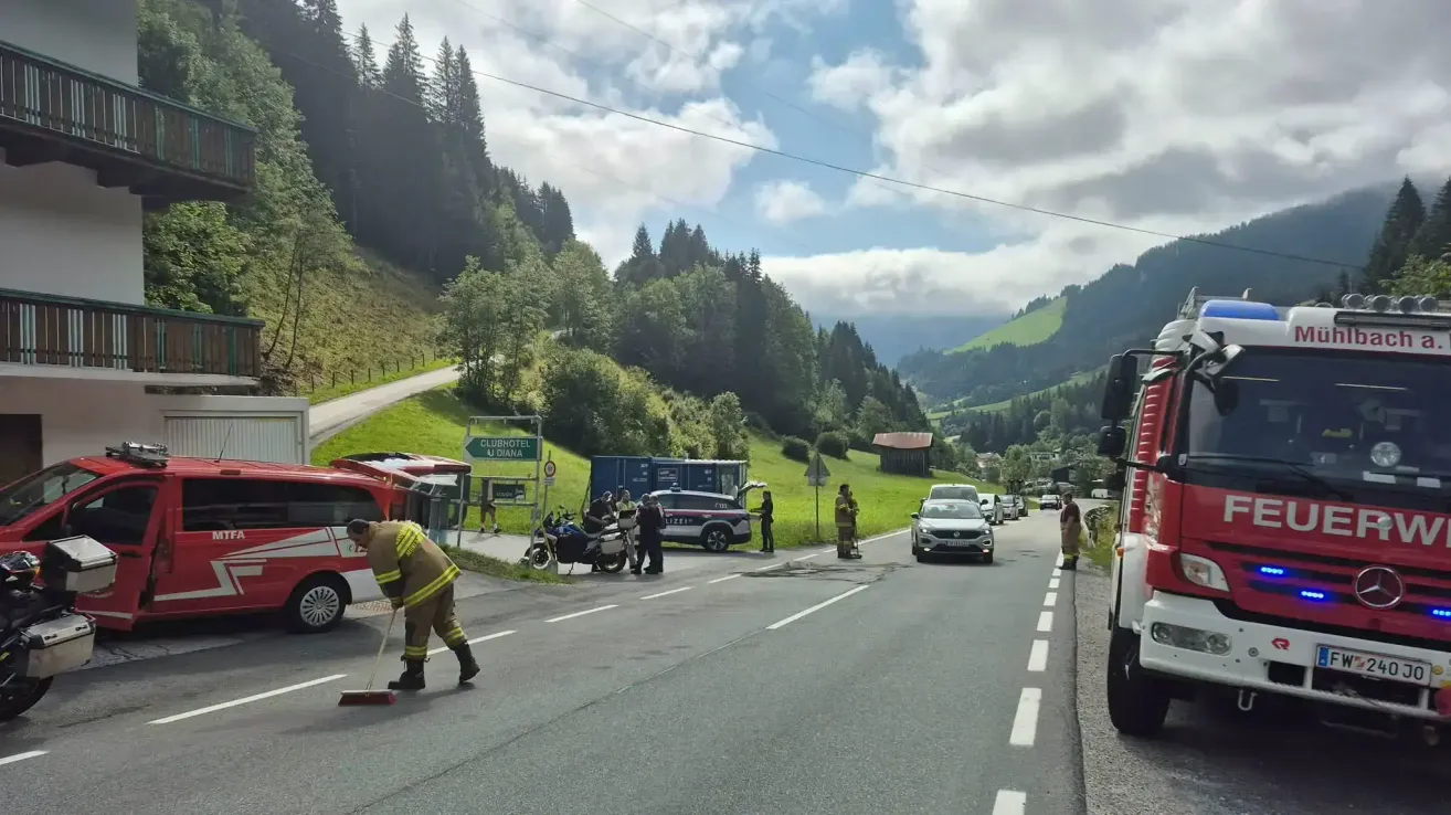 Ein Feuerwehrmann fegt die Straße, während Rettungsfahrzeuge am Straßenrand geparkt sind. Ein Motorrad und Autos sind auf der Straße angehalten. Bäume und Berge sind im Hintergrund.