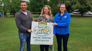Drei Personen stehen in einem Park und halten ein Schild mit der Aufschrift 'Natur im Garten Kindergarten'. Die Frau in der Mitte trägt eine Camouflage-Bluse und eine Brille. Hinter ihnen befindet sich ein Spielplatz mit einer Schaukel.