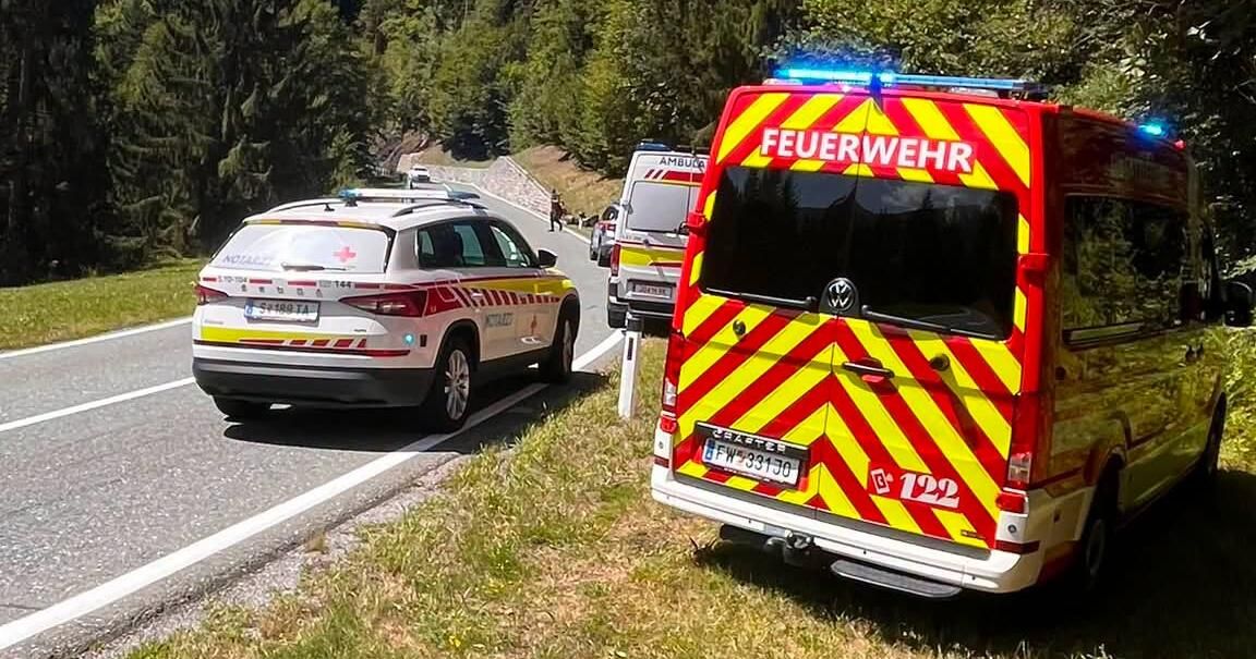 Rettungsfahrzeuge, darunter ein Feuerwehrwagen und ein Krankenwagen, sind am Straßenrand geparkt, mit Bäumen und einem Berg im Hintergrund.