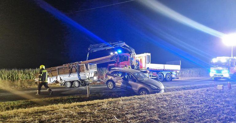Ein Auto ist nachts an der Straßenseite demoliert. Ein Kran hebt ein Stück Holz vom Auto. Ein Feuerwehrwagen ist hinter dem Kran geparkt.