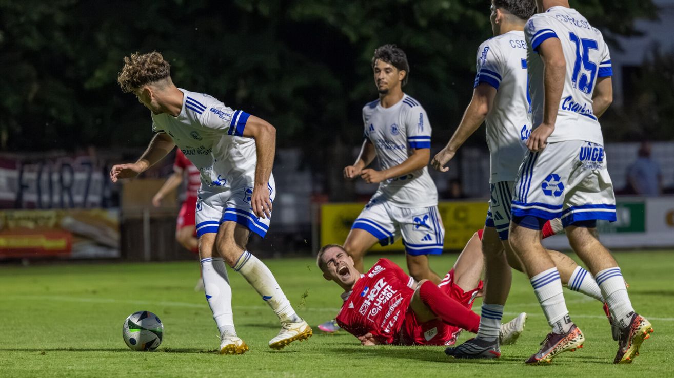 Ein Fußballspieler in einem weißen Trikot läuft auf einen gefallenen Spieler in einem roten Trikot auf einem Feld zu.