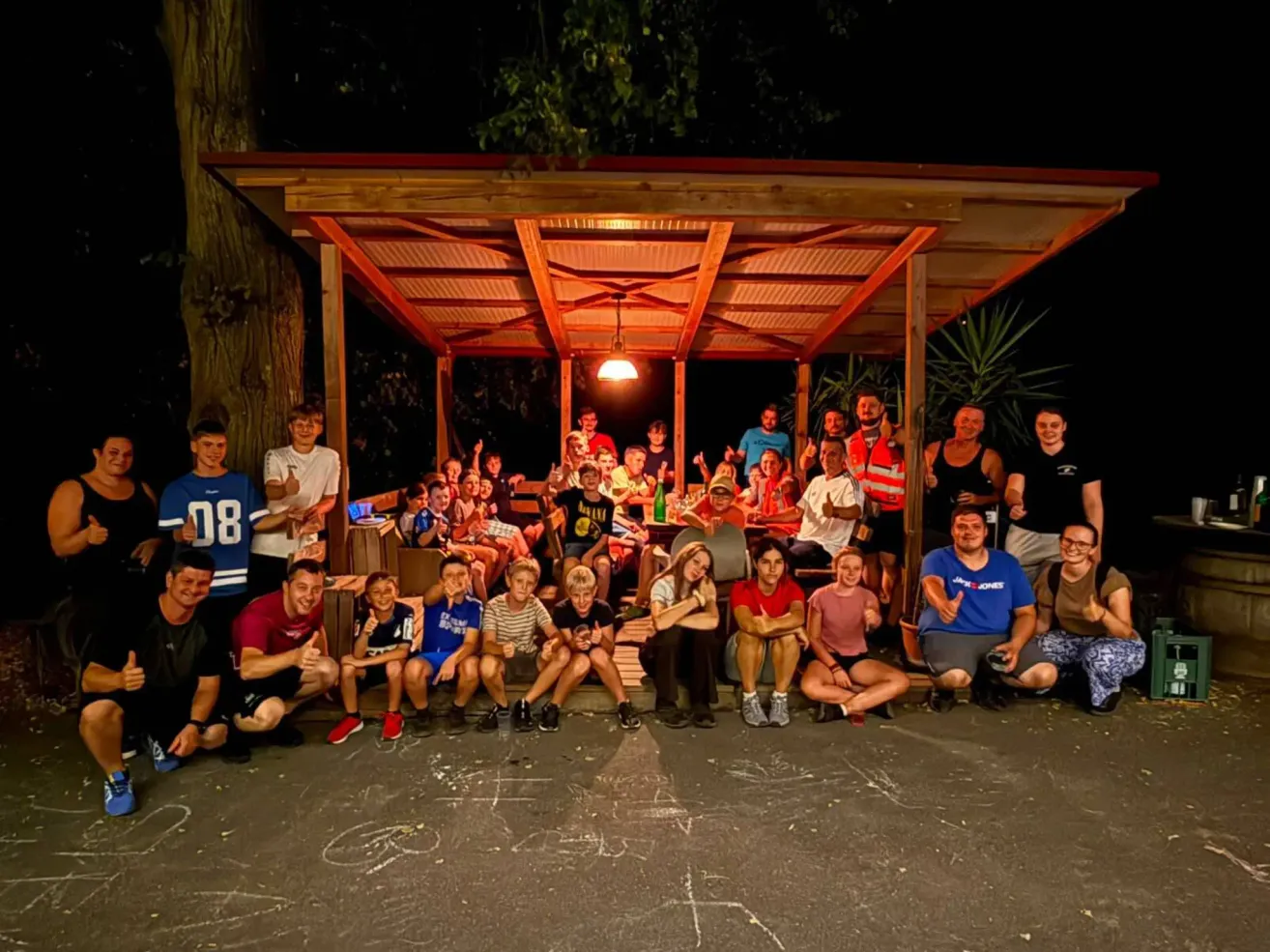 Eine Gruppe von Menschen versammelt sich nachts unter einem Pavillon, einige sitzen und andere stehen, wobei einige Personen Getränke in der Hand halten. Die Umgebung ist im Freien mit einem Baum und Pflanzen im Hintergrund.
