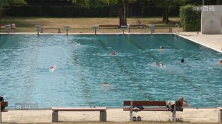 Eine Gruppe von Menschen schwimmt in einem großen Freibad. Das Becken ist von Bänken und Sprungbrettern umgeben.
