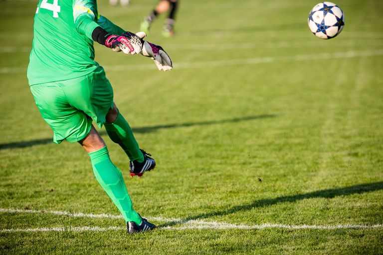 Ein Fußballtorwart in grüner Uniform, Handschuhen und Stollenschuhen, der springt, um einen Ball zu fangen. Das Feld ist grün mit weißen Markierungen.
