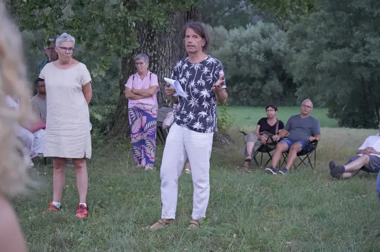 Eine Gruppe von Menschen steht auf einer Rasenfläche. Ein Mann in der Mitte hält ein Papier und spricht. Hinter ihm steht eine Frau mit gefalteten Händen. Einige Leute sitzen auf Stühlen. Ein Baum steht hinter ihnen.