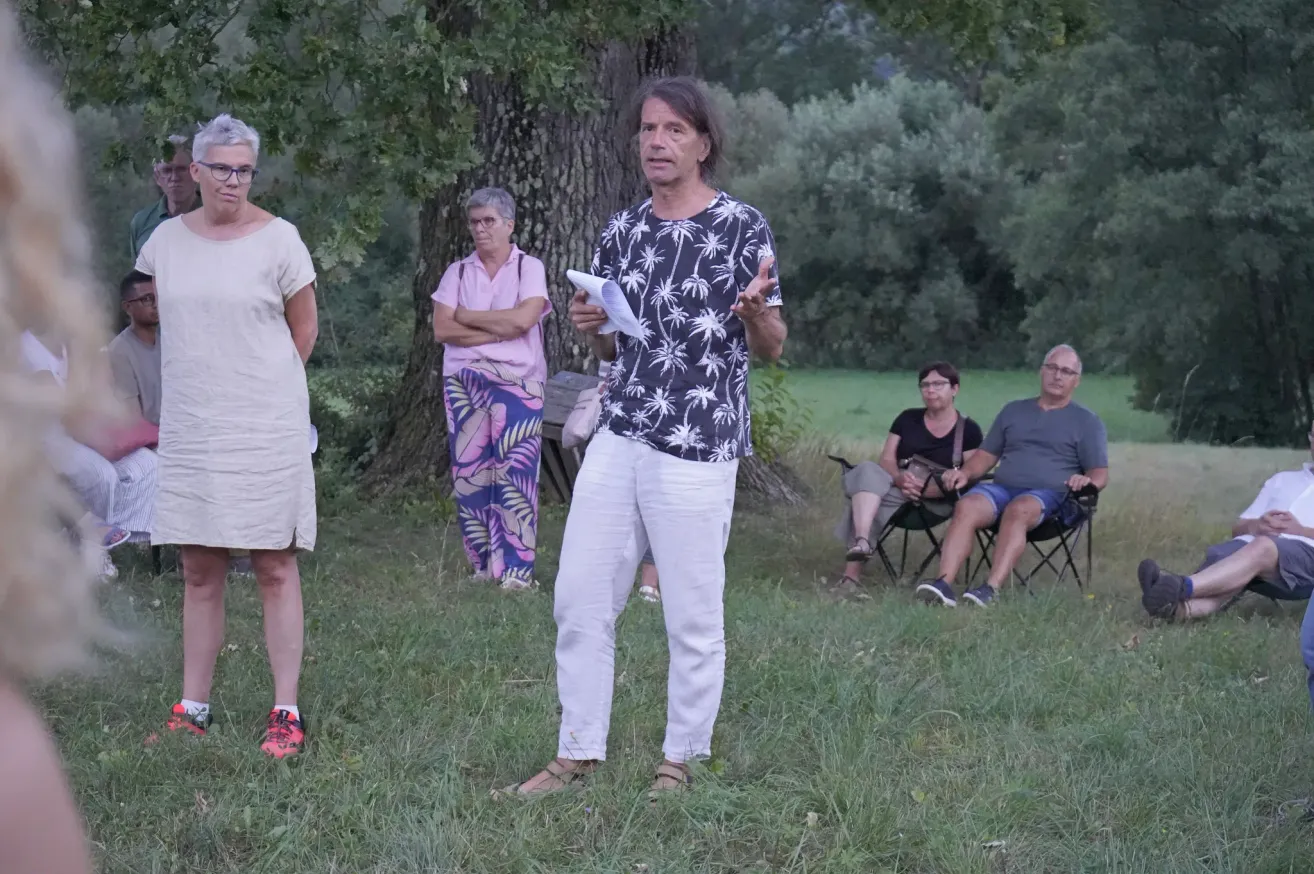 Eine Gruppe von Menschen steht auf einer Rasenfläche. Ein Mann in der Mitte hält ein Papier und spricht. Hinter ihm steht eine Frau mit gefalteten Händen. Einige Leute sitzen auf Stühlen. Ein Baum steht hinter ihnen.
