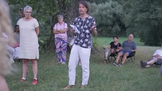 Eine Gruppe von Menschen steht auf einer Rasenfläche. Ein Mann in der Mitte hält ein Papier und spricht. Hinter ihm steht eine Frau mit gefalteten Händen. Einige Leute sitzen auf Stühlen. Ein Baum steht hinter ihnen.